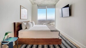 Ein Schlafzimmer mit einem großen Bett, einem Schreibtisch, einem Sessel, einem an der Wand montierten Fernseher und einem Fenster mit Blick auf die Stadt.