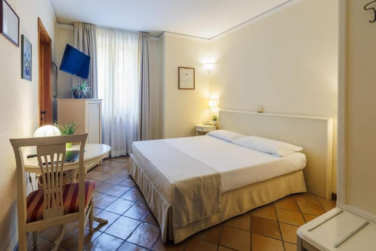 A hotel room with a bed, a desk with a chair, a TV, a lamp, and a window with curtains.