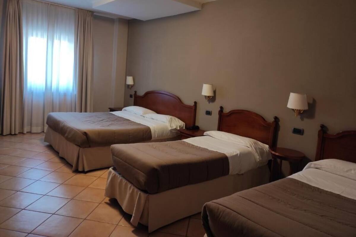 A hotel room with three beds, a window with curtains, and two wall-mounted lamps.