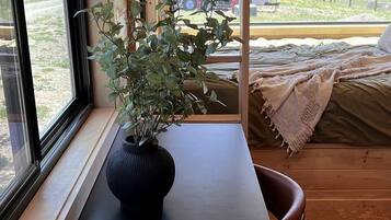A wooden interior with a table, a potted plant, and a bed with a view of a rural landscape.