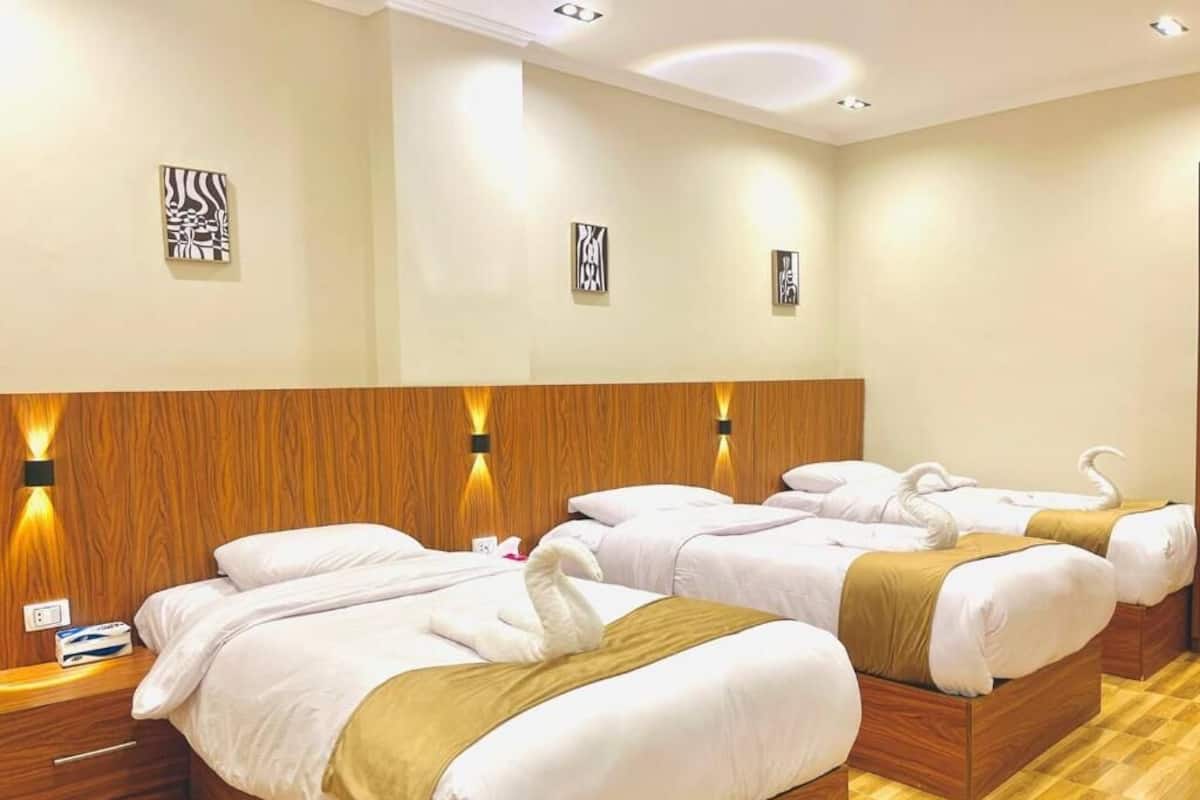 Quarto de hotel com quatro camas, cabeceiras de madeira e luminárias fixadas na parede.
