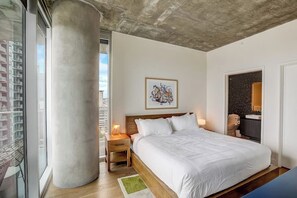 A bedroom with a large window, a bed, a nightstand, a framed picture, and a bathroom door. - 16th floor condo w/rooftop pool & walkable location (Austin)