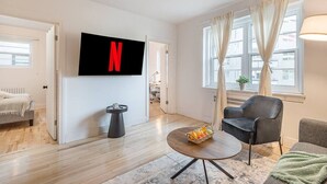 Una sala moderna con una televisión de pantalla plana, una mesa redonda con fruta, un sofá gris y una cama al fondo.