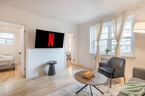 Ein modernes Wohnzimmer mit einem Flachbildfernseher, einem runden Couchtisch mit Früchten, einem grauen Sofa und einem Bett im Hintergrund.
