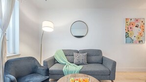 Una sala moderna con un sofá gris, un sillón negro, una mesa redonda de centro con fruta, una lámpara de pie y un espejo fijado a la pared.
