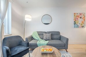 Ein modernes Wohnzimmer mit einem grauen Sofa, einem schwarzen Sessel, einem runden Couchtisch mit Obst, einer Stehlampe und einem wandmontierten Spiegel.