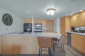 A kitchen with a central island, bar stools, wooden cabinets, a black refrigerator, an oven, a microwave, and a clock on the wall.