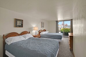 A hotel room with two beds, a nightstand, a lamp, and a window with a view of buildings.