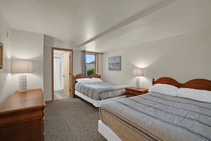 A bedroom with two beds, a nightstand, a lamp, a door leading to another room, and a window with a view.