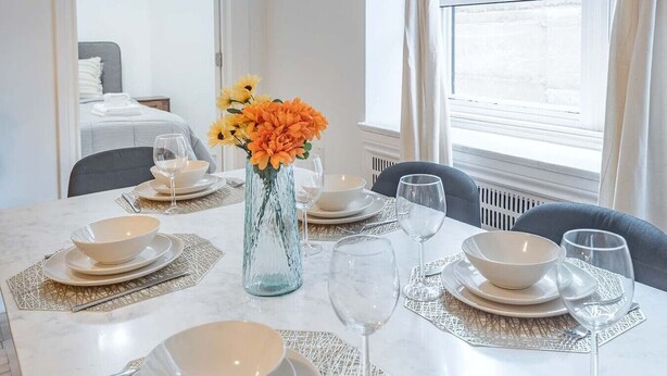 Ein Esstisch ist gedeckt mit weißen Tellern, Schüsseln und Weingläsern, eine Vase mit orangefarbenen Blumen, und im Hintergrund sieht man ein Bett und ein Fenster.
