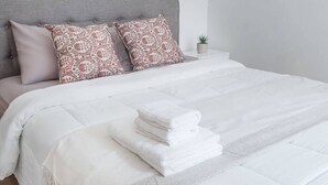 Ein ordentlich bezogenes Bett mit weißer Decke und gefaltetem weißen Handtuch, ein grauer Stoffkopfteil und zwei dekorative Kissen.