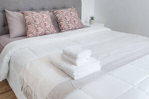 Ein ordentlich bezogenes Bett mit weißer Decke und gefaltetem weißen Handtuch, ein grauer Stoffkopfteil und zwei dekorative Kissen.
