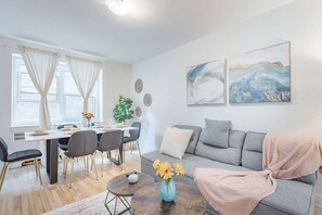 Ein modernes Wohnzimmer mit einem grauen Sofa, einem Esstisch mit Stühlen und zwei abstrakten Bildern an der Wand.