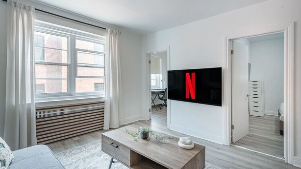 A modern living room with a large TV mounted on the wall, a window with curtains, a radiator, and a wooden coffee table. - Spacious Renovated 3BR | Free Parking (Montreal)