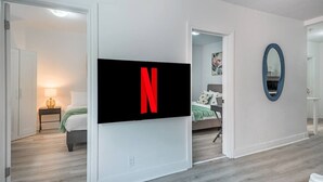 A modern hotel room with a large flat-screen TV mounted on the wall, a bed with a green blanket, a bedside table with a lamp, and a mirror on the wall. - Spacious Renovated 3BR | Free Parking (Montreal)
