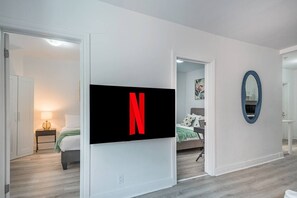 Ein modernes Hotelzimmer mit einem großen Flachbildfernseher an der Wand, einem Bett mit grüner Decke, einem Nachttisch mit Lampe und einem Spiegel.