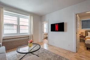 Ein modernes Hotelzimmer mit großem Fenster, einem runden Glastisch mit Vase, einem Flachbildfernseher an der Wand und einem Bett mit Nachttisch.
