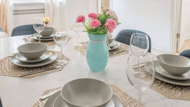 Una mesa de comedor dispuesta con platos, tazones y copas de vino, junto con un jarrón de flores.
