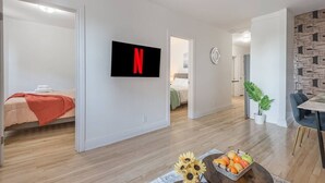 Un apartamento moderno con un televisor de pantalla plana, una zona de comedor con mesa y sillas, una cama con una colcha roja y un pasillo que conduce a otra habitación.