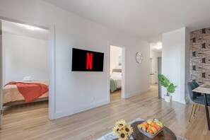 A modern apartment with a flat-screen TV, a dining area with a table and chairs, a bed with a red blanket, and a hallway leading to another room. - Elegant 3BR- Concordia- Indoor Parking / AC (Montréal)
