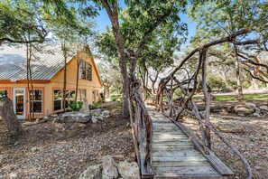 Eine rustikale Holzbrücke führt zu einem Haus mit geneigtem Dach, umgeben von Bäumen und einem Kiesweg.