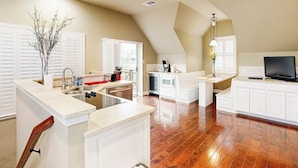 A kitchen with white cabinetry, a central island with a sink, and a dining area with a TV. - Countryside 3-story home with game room, WFH office & guest house with kitchen (Volente)