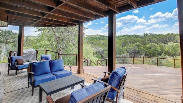 A covered outdoor deck with seating and a table, overlooking a natural landscape. - Countryside 3-story home with game room, WFH office & guest house with kitchen (Volente)