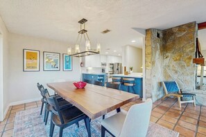 Ein Essbereich mit Holztisch, Stühlen und einer Chandelier. Im Hintergrund ist eine Küche mit blauen Schränken und einer Wand mit Steinverkleidung zu sehen.