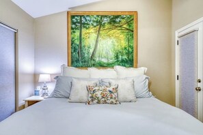 Ein Schlafzimmer mit Bett, Kissen, einem Nachttisch, einer Lampe und einem Waldgemälde.