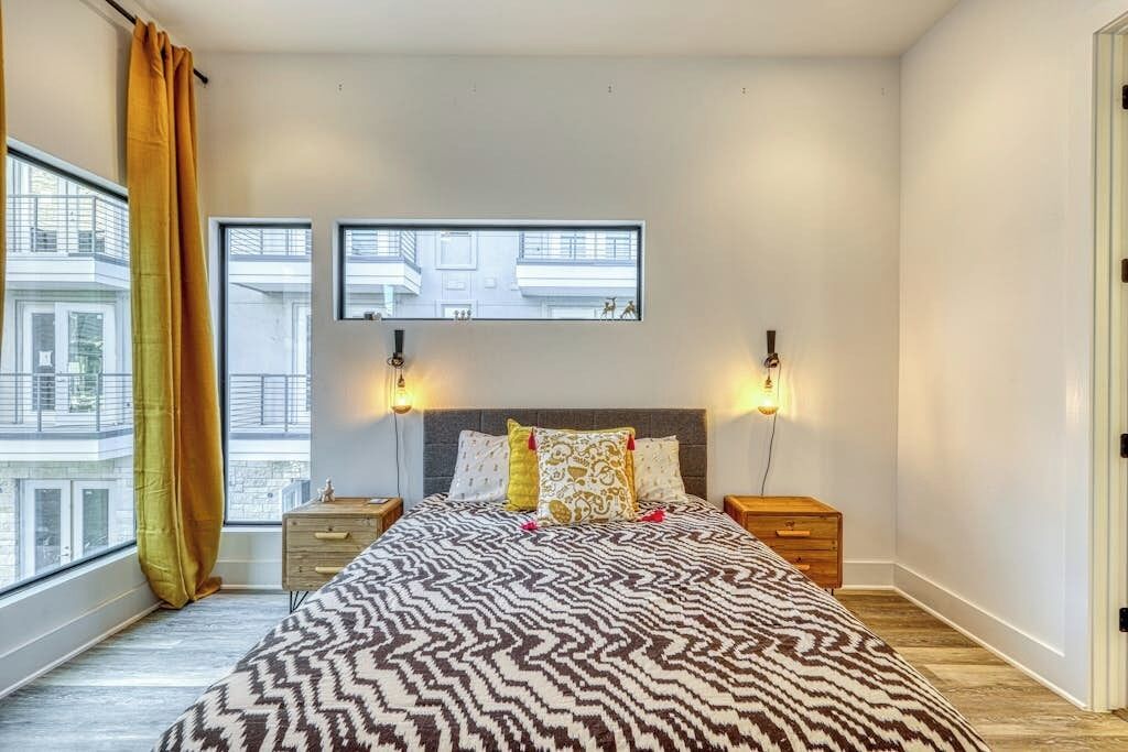 A bedroom with a bed, two bedside tables, a window with curtains, and a wall-mounted TV.