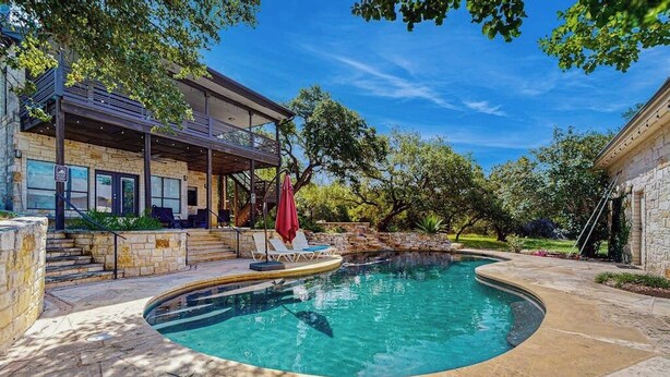 A swimming pool with a curved edge, surrounded by stone tiles and a covered patio area with outdoor furniture. - Contemporary dog-friendly house w/ private gas grill, hot tub, & outdoor pool (Austin)