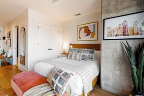 Ein Schlafzimmer mit einem Bett, Nachttischen, einem Spiegel, einem Stadtbild im Bilderrahmen und einer Topfpflanze.