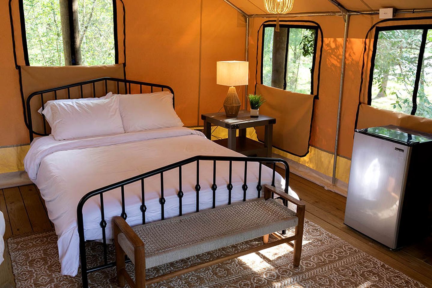 A bed with a metal frame, a small bedside table with a lamp, a bench, and a refrigerator.
