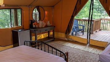 A tent with a bed, a dresser, and a chair on a deck with a view of trees.
