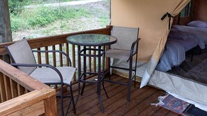 A cozy outdoor deck with a table and chairs, a tent with a bed inside, and a view of the surrounding forest. - Great Small Group Safari Tent in Livingston, Texas (Livingston)