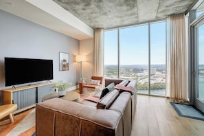 Ein modernes Wohnzimmer mit einem großen Flachbildfernseher, einer Couch, einem Couchtisch und Blick auf eine Skyline durch große Fenster.