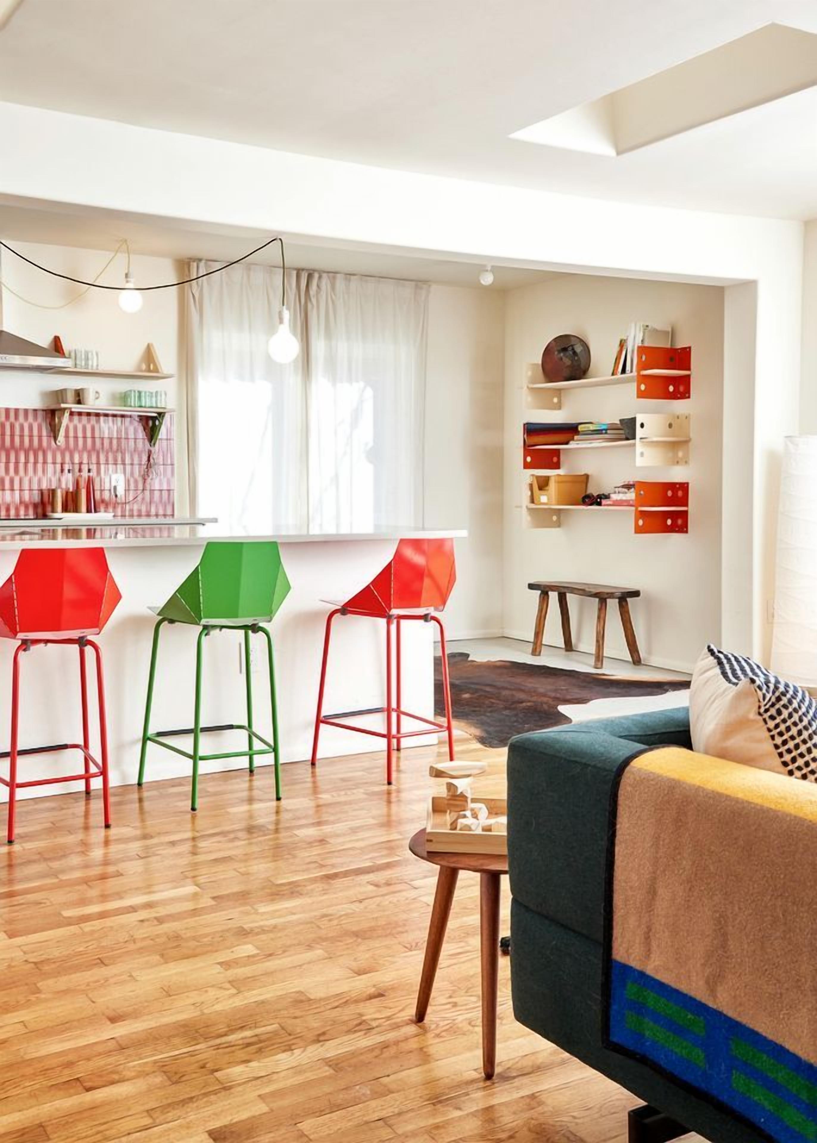 A modern living space with a kitchen area, wooden flooring, and colorful stools.