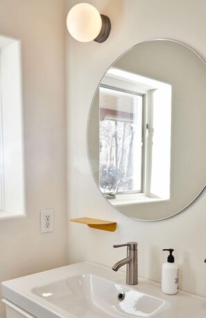 A bathroom with a white sink, a round mirror, a wall-mounted light, and a window.