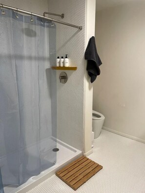 A shower with a transparent curtain, a wooden shelf with toiletries, a towel rack, and a white toilet.