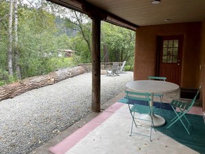 An outdoor patio with a round table and chairs, a wooden door, and a view of a forested area.