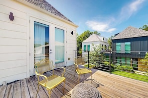 Eine Holzterrasse mit Sitzgelegenheiten, ein weißes Gebäude mit Glastür und ein modernes Haus im Hintergrund.