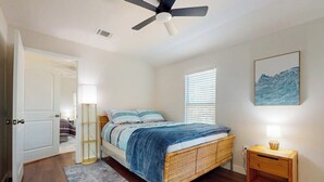 A bedroom with a bed, a ceiling fan, a bedside table with a lamp, a window with blinds, and a wall-mounted artwork. - Modern cozy house with full kitchen, dining & living area, TV, close to downtown (Austin)