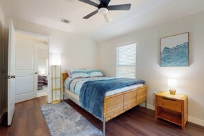 Ein Schlafzimmer mit Bett, Deckenventilator, Nachttisch mit Lampe, Fenster mit Jalousien und Wandbild.