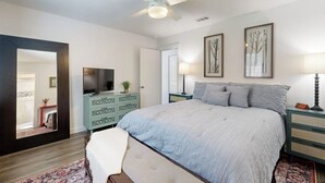 Un dormitorio con cama, ventilador de techo, televisión, cómoda y cuadros en la pared.