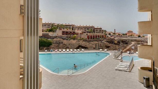 A swimming pool with lounge chairs, a view of a building with multiple floors, and a rocky landscape in the background. - Charming 2-bedroom Apartment With Ocean Views in Adeje (Adeje)