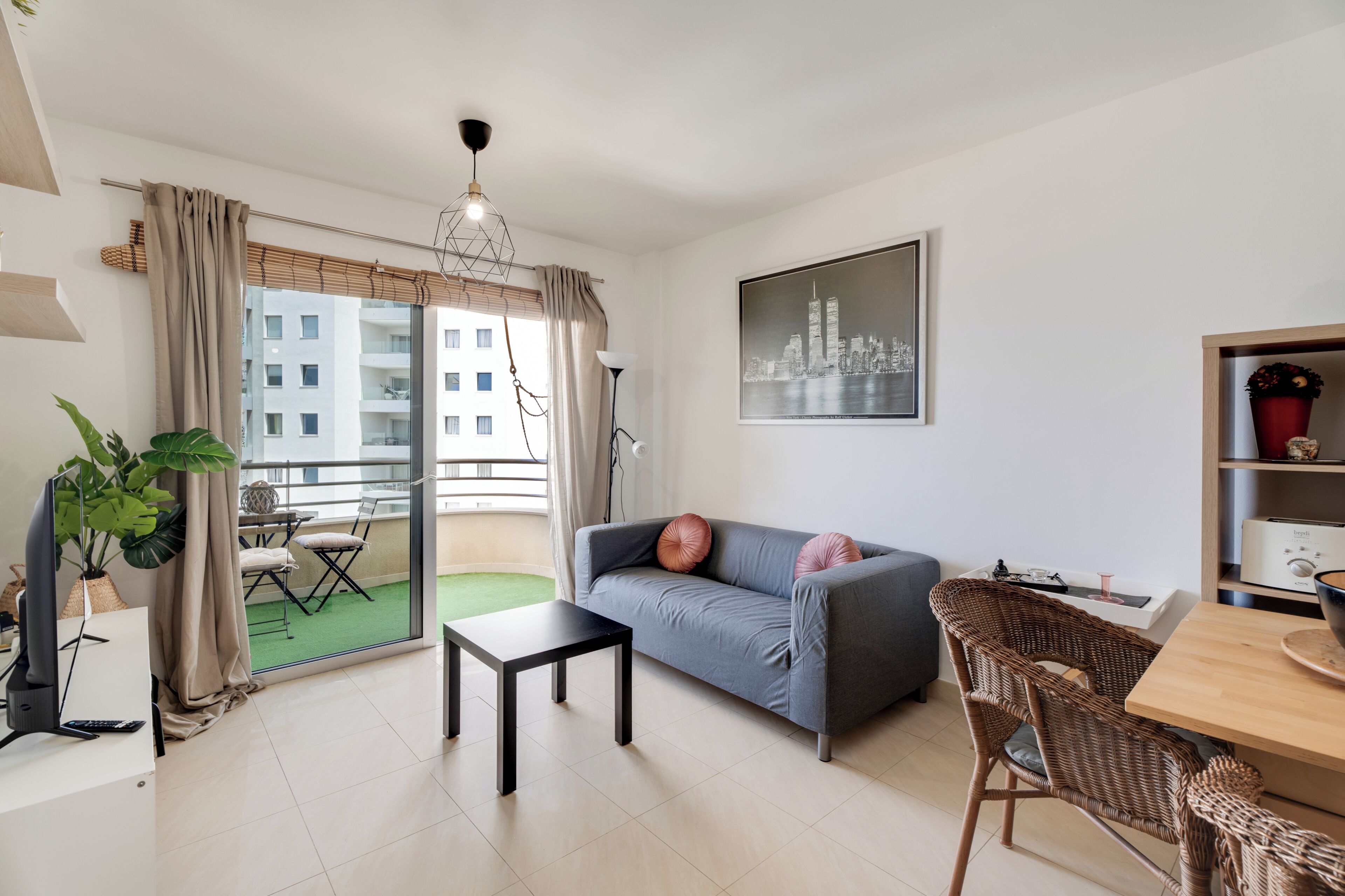 Un salón moderno con un sofá, una mesa de comedor, una mesita, una silla, una planta en maceta y un balcón con vistas a edificios.