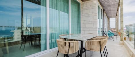 Un área de comedor exterior moderna con pared de vidrio, pérgola de madera y vista al mar.