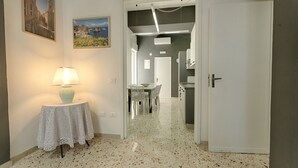 Een gang met een tafel en lamp, een zithoek met stoelen, en een keuken met kasten en een deur.