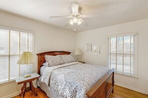 Ein Schlafzimmer mit Bett, Nachttisch, Lampe, Deckenventilator und zwei Fenstern mit Jalousien.