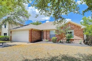 A single-story house with a garage, brick exterior, and a small front yard with trees and shrubs.